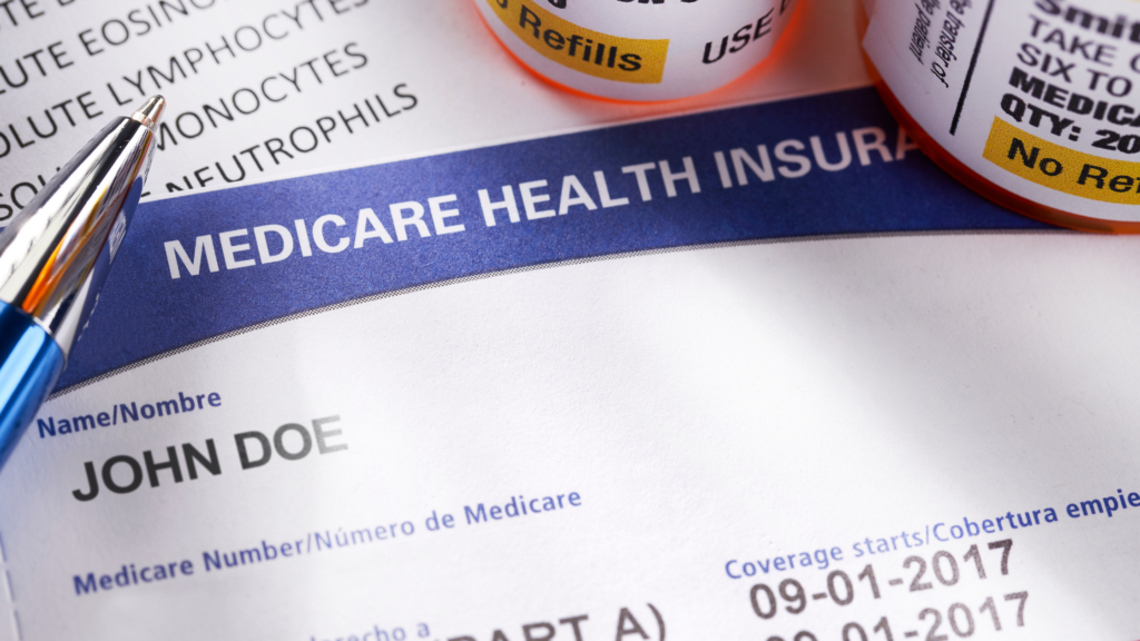 This is an image of a Medicare health insurance card lying on a surface next to a pen and a prescription bottle. The card has the name "John Doe" and other details like the Medicare number and coverage dates, indicating it is a mock-up or sample card often used for illustrative purposes. The prescription bottle label in the background and the pen suggest a healthcare or medical setting.